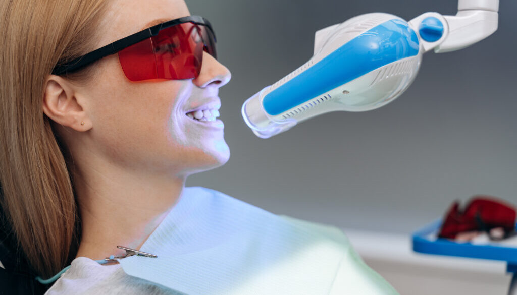 Attractive, young patient in a dental chair with goggles shows her teeth. Dental equipment shines on the patient's teeth